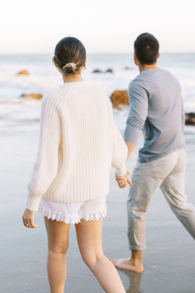A woman and man holding hands, walking barefoot along the beach. The woman wears a white sweater and shorts, while the man wears a light blue shirt and light pants. The ocean and rocks are visible in the background. Copyright - Lisa Leanne Photography, California