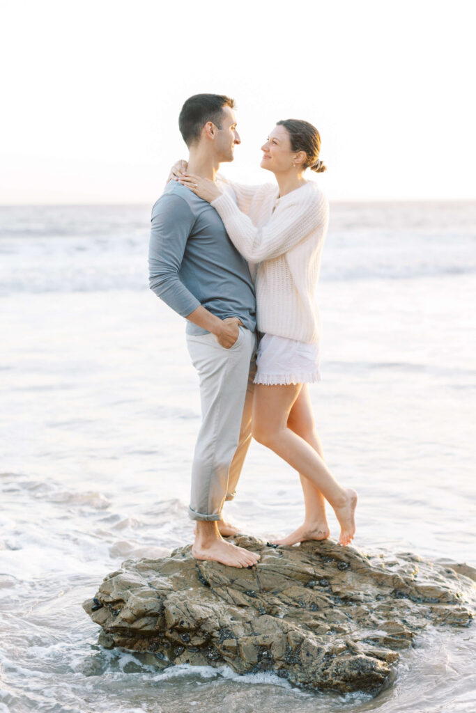 A couple stands barefoot on a rock at the beach, smiling and looking into each other's eyes. The woman has her arms around the man's neck, and the ocean waves surround them under a bright sky. Copyright - Lisa Leanne Photography, California