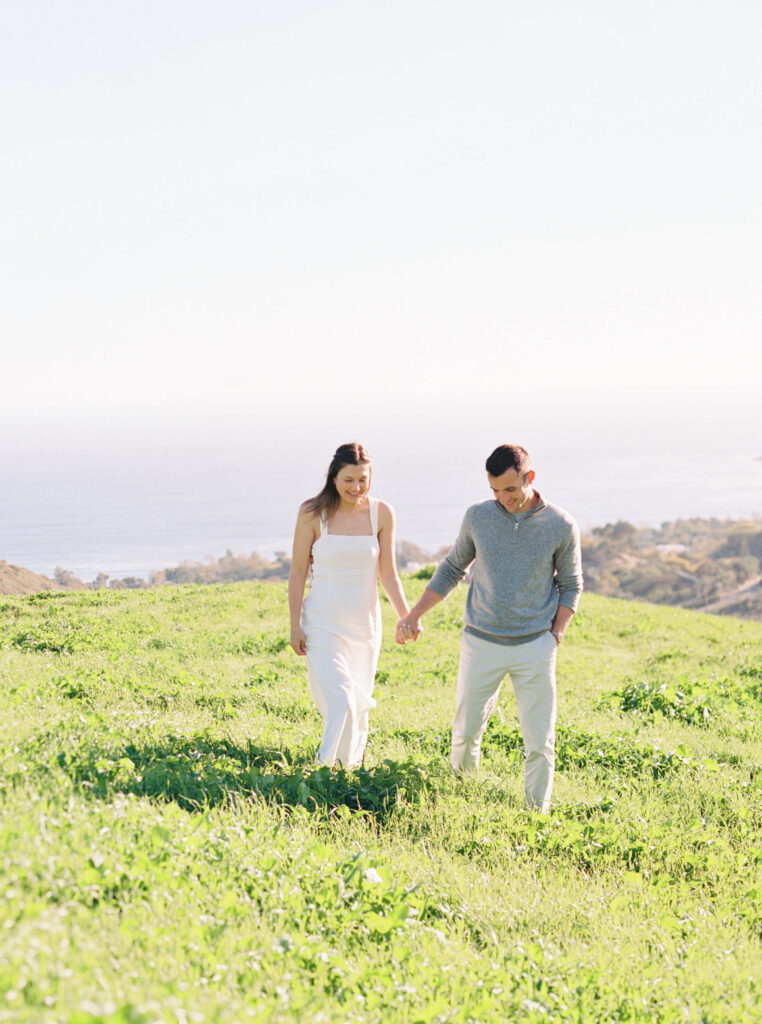 A couple walks hand in hand through a green grassy field under a clear sky, with the ocean and coastline visible in the background. Both are dressed in light, casual clothing and appear happy and relaxed. Copyright - Lisa Leanne Photography, California