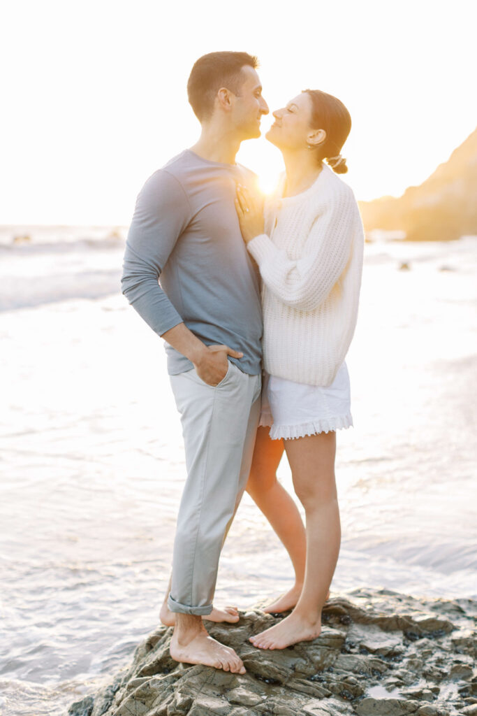 A couple stands barefoot on a rocky beach at sunset, facing each other and smiling. The woman wears a white sweater and shorts, while the man wears a light blue shirt and pants. Sunlight softly illuminates them. Copyright - Lisa Leanne Photography, California