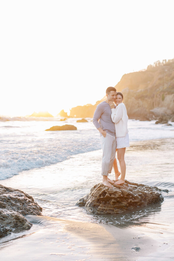 A couple stands barefoot on a rock at the beach during sunset, smiling and embracing as gentle waves wash around them. The sky is bright and the scene feels warm and romantic. Copyright - Lisa Leanne Photography, California