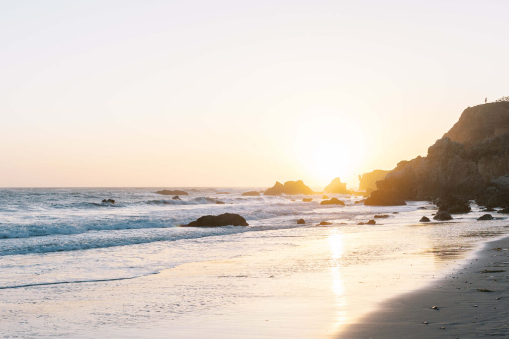 Sunset over a rocky beach with waves gently washing onto the shore, sunlight reflecting on the water and sand, and a cliffside visible on the right. Copyright - Lisa Leanne Photography, California