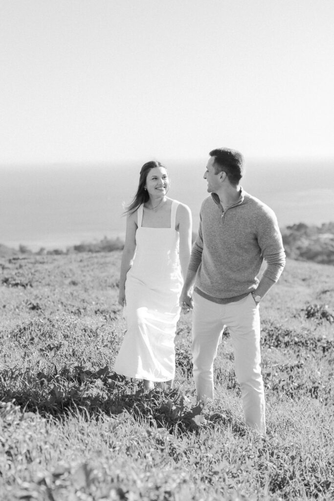 A woman in a white dress and a man in a sweater and light pants walk together through a grassy field, smiling at each other with the ocean visible in the background. The image is black and white. Copyright - Lisa Leanne Photography, California