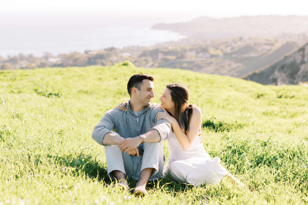 A couple sits together on green grass, smiling at each other, with rolling hills and a hazy coastline in the background on a sunny day. Copyright - Lisa Leanne Photography, California