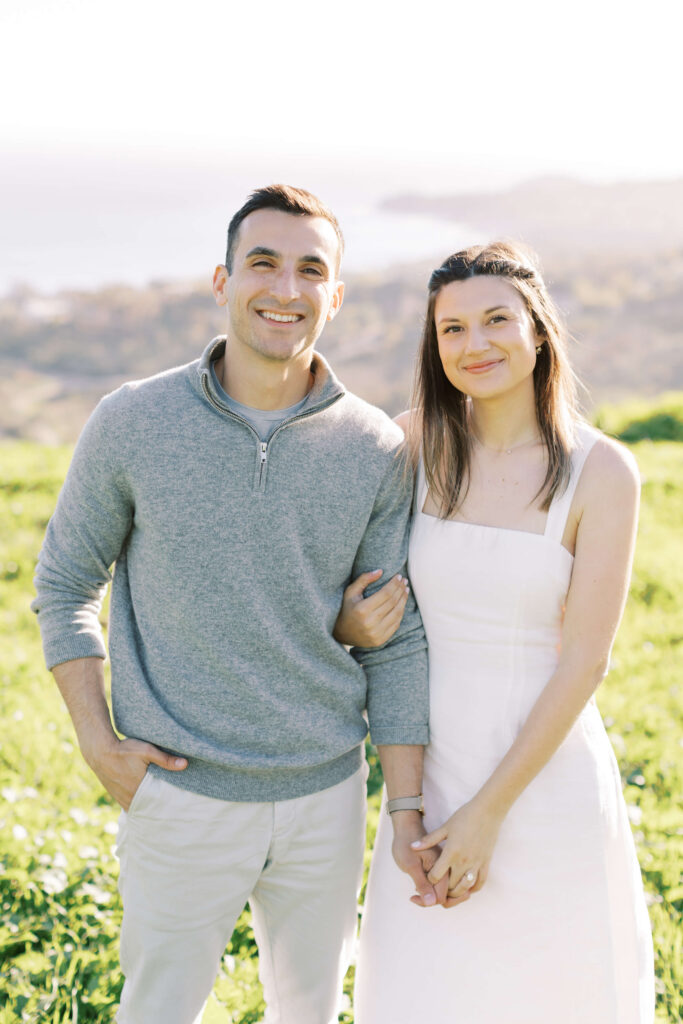 A smiling couple stands outdoors in a sunlit field. The man wears a light gray sweater and pants, and the woman wears a sleeveless white dress. They are standing close together, arm in arm, with a blurred landscape in the background. Copyright - Lisa Leanne Photography, California