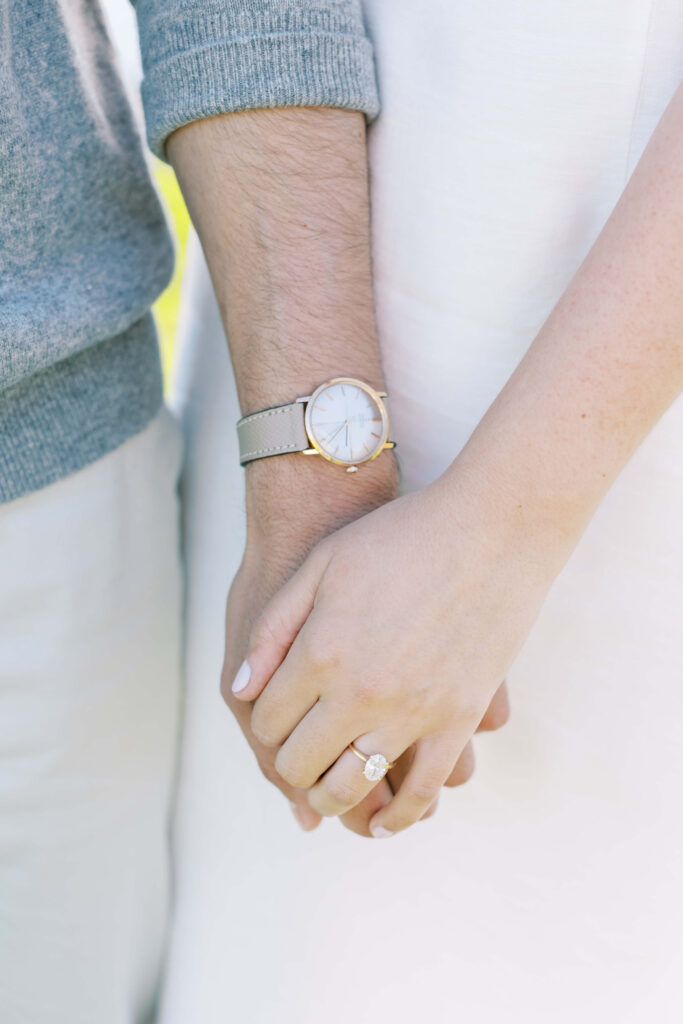 Close-up of a couple holding hands. One person wears a watch and a gray sweater; the other wears a white dress and an engagement ring. Copyright - Lisa Leanne Photography, California