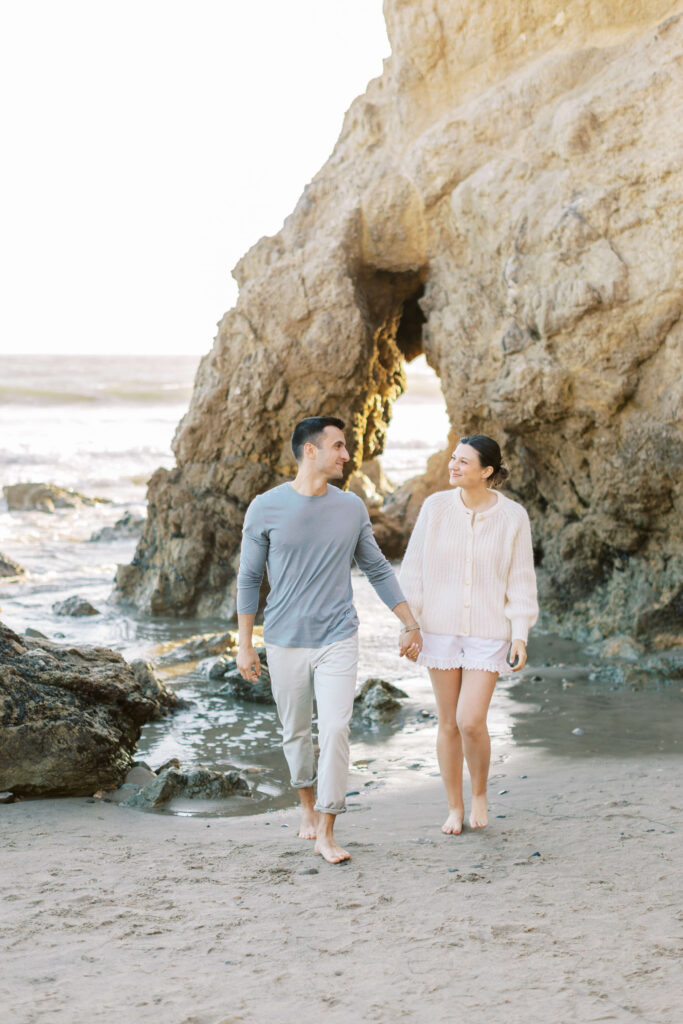 A couple walks barefoot on a sandy beach, holding hands and smiling at each other, with large rocky formations and the ocean in the background. Copyright - Lisa Leanne Photography, California