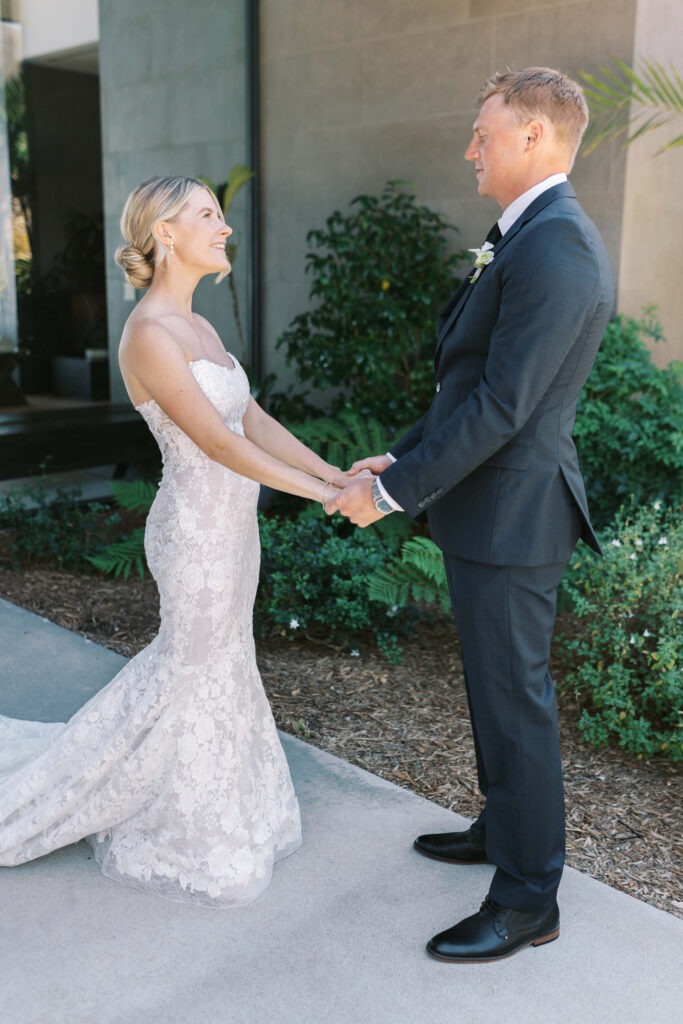 Malibu-Estate-Wedding_Malibu-Wedding_Southern-California-Wedding-Photographer_Lisa-Leanne-Photography_10-683x1024 A bride in a white lace gown and a groom in a dark suit stand facing each other, holding hands and smiling outdoors near greenery and a building. Copyright - Lisa Leanne Photography, California