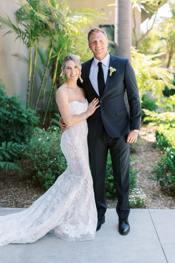 Malibu-Estate-Wedding_Malibu-Wedding_Southern-California-Wedding-Photographer_Lisa-Leanne-Photography_12-683x1024 A bride in a strapless lace wedding dress and a groom in a dark suit with a boutonnière pose together, smiling outdoors amid green plants and trees. Copyright - Lisa Leanne Photography, California