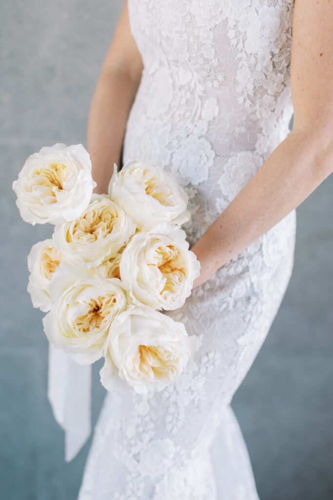 Malibu-Estate-Wedding_Malibu-Wedding_Southern-California-Wedding-Photographer_Lisa-Leanne-Photography_13-683x1024 A person in a white lace dress holds a bouquet of large, creamy white roses with soft yellow centers, standing against a light gray background. Copyright - Lisa Leanne Photography, California