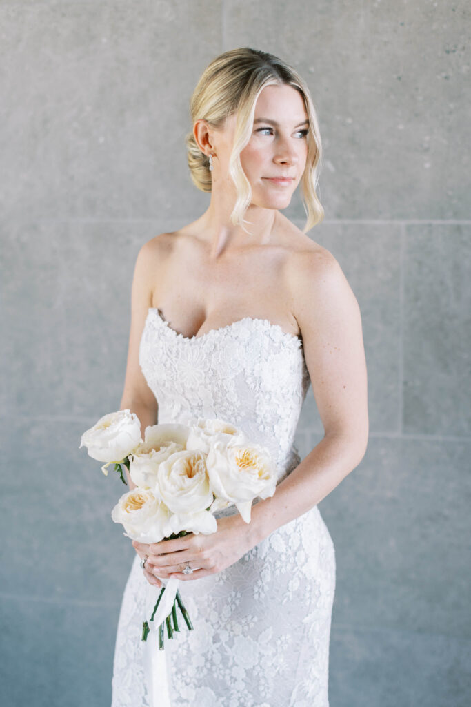Malibu-Estate-Wedding_Malibu-Wedding_Southern-California-Wedding-Photographer_Lisa-Leanne-Photography_15-683x1024 A bride in a strapless white lace gown holds a bouquet of white roses, standing against a light gray stone wall with her blonde hair styled in a low updo and loose tendrils framing her face. Copyright - Lisa Leanne Photography, California