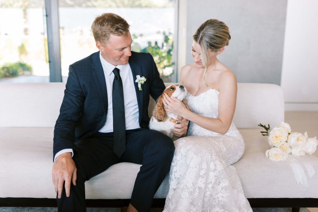 Malibu-Estate-Wedding_Malibu-Wedding_Southern-California-Wedding-Photographer_Lisa-Leanne-Photography_16-1024x683 A bride in a white lace gown and a groom in a dark suit sit on a light sofa, smiling and holding a small brown and white dog. A bouquet of white flowers rests beside the bride. Copyright - Lisa Leanne Photography, California