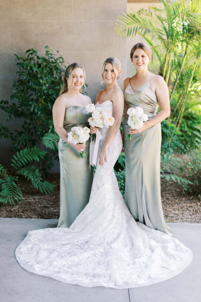 Malibu-Estate-Wedding_Malibu-Wedding_Southern-California-Wedding-Photographer_Lisa-Leanne-Photography_17-683x1024 Three women posing outdoors; the bride stands in the center wearing a white lace gown and holding a bouquet, flanked by two bridesmaids in sage green dresses, also holding white bouquets. Green foliage is in the background. Copyright - Lisa Leanne Photography, California