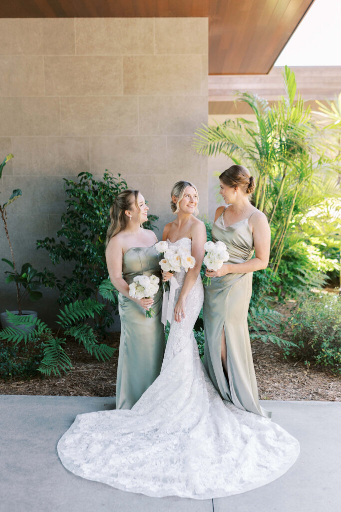 Malibu-Estate-Wedding_Malibu-Wedding_Southern-California-Wedding-Photographer_Lisa-Leanne-Photography_18-683x1024 Three women in formal dresses pose outside. The woman in the center wears a white lace wedding gown and holds white flowers. The two women beside her wear matching light green dresses and also hold bouquets, all smiling together. Copyright - Lisa Leanne Photography, California