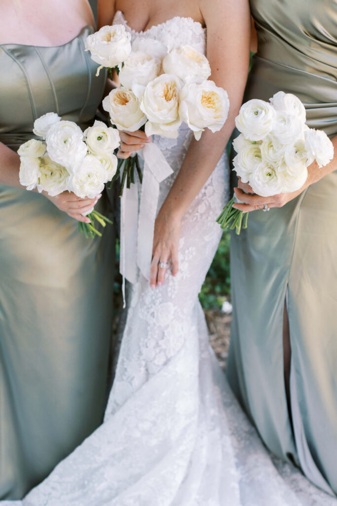 Malibu-Estate-Wedding_Malibu-Wedding_Southern-California-Wedding-Photographer_Lisa-Leanne-Photography_19-683x1024 Three women in formal dresses hold bouquets of white flowers. The central figure wears a white lace wedding gown, while the women on either side wear sage green dresses. The image is cropped to show them from shoulders down. Copyright - Lisa Leanne Photography, California