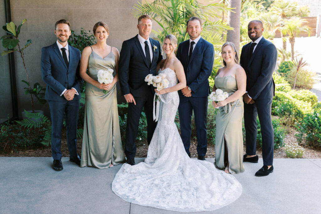 Malibu-Estate-Wedding_Malibu-Wedding_Southern-California-Wedding-Photographer_Lisa-Leanne-Photography_20-1024x683 A bride and groom stand together outside, flanked by three bridesmaids in light green dresses holding white bouquets and two groomsmen in dark suits, all smiling at the camera with greenery in the background. Copyright - Lisa Leanne Photography, California