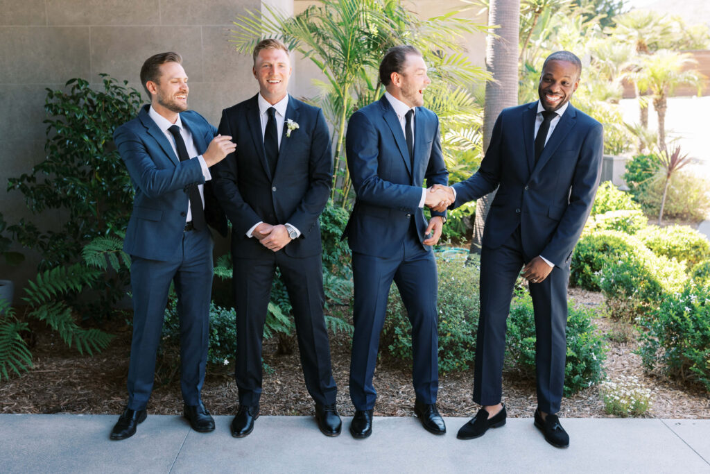 Malibu-Estate-Wedding_Malibu-Wedding_Southern-California-Wedding-Photographer_Lisa-Leanne-Photography_21-1024x683 Four men in suits stand outside in front of greenery. Two are laughing while one shakes hands with another, and the fourth smiles with his hands by his side. It appears to be a lighthearted moment at a formal event. Copyright - Lisa Leanne Photography, California