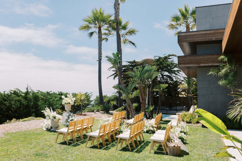 Malibu-Estate-Wedding_Malibu-Wedding_Southern-California-Wedding-Photographer_Lisa-Leanne-Photography_23-1024x683 An outdoor wedding ceremony setup with wooden chairs arranged on grass, surrounded by tropical plants and palm trees, white floral decorations, and a modern building to the right under a blue sky. Copyright - Lisa Leanne Photography, California
