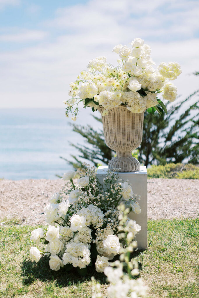 Malibu-Estate-Wedding_Malibu-Wedding_Southern-California-Wedding-Photographer_Lisa-Leanne-Photography_25-683x1024 A white wicker vase filled with white roses and flowers sits on a pedestal outdoors by the sea, with more white flowers arranged at its base on green grass under a bright sky. Copyright - Lisa Leanne Photography, California