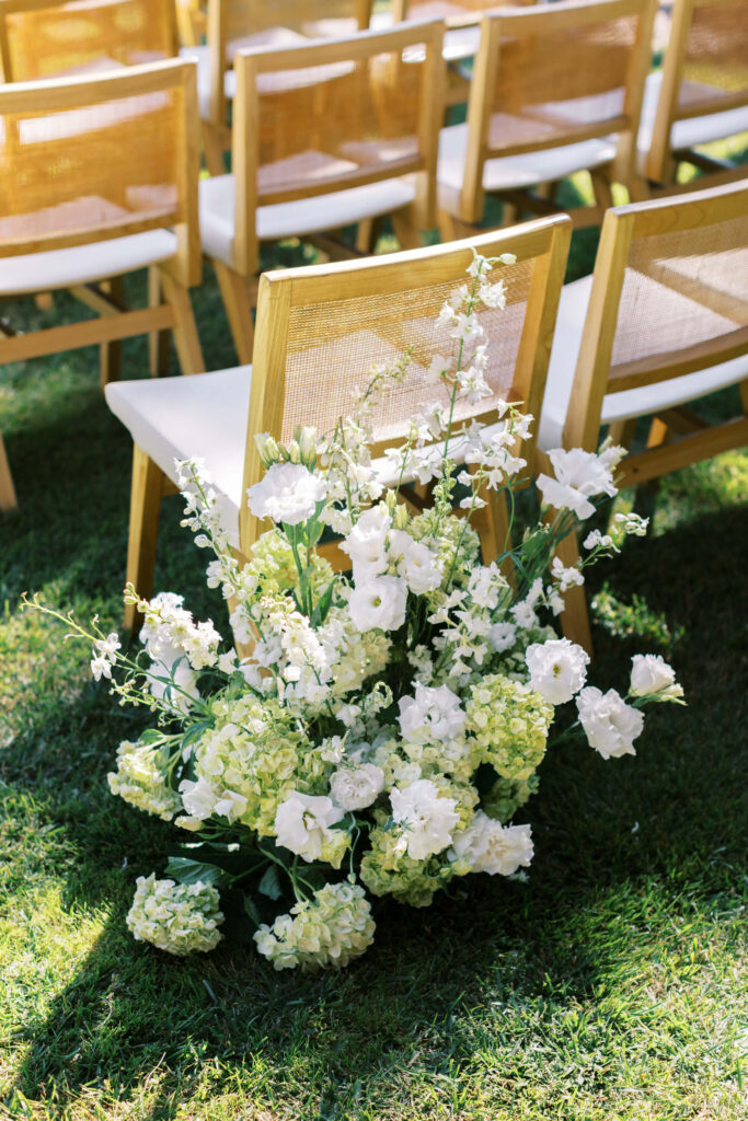 Malibu-Estate-Wedding_Malibu-Wedding_Southern-California-Wedding-Photographer_Lisa-Leanne-Photography_26-683x1024 Rows of wooden chairs with white cushions are arranged outdoors on grass. A large floral arrangement with white and light yellow flowers decorates the aisle beside the chairs. Copyright - Lisa Leanne Photography, California
