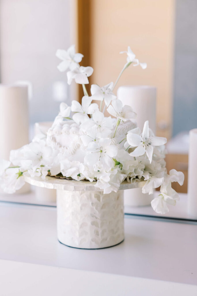 Malibu-Estate-Wedding_Malibu-Wedding_Southern-California-Wedding-Photographer_Lisa-Leanne-Photography_27-683x1024 A small white cake decorated with white flowers sits on a textured white cake stand. The background features blurred cylindrical objects and a soft, neutral setting. Copyright - Lisa Leanne Photography, California
