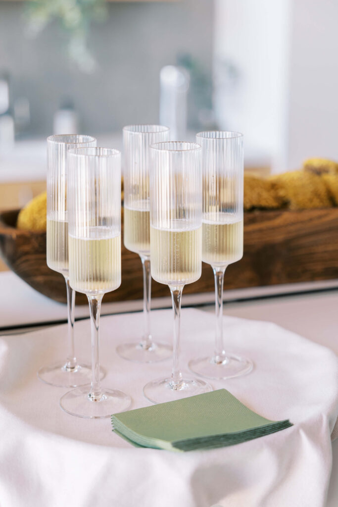 Malibu-Estate-Wedding_Malibu-Wedding_Southern-California-Wedding-Photographer_Lisa-Leanne-Photography_29-683x1024 Six tall champagne flutes filled with sparkling wine are arranged on a round white tablecloth, with a stack of green napkins in the foreground and a wooden bowl with moss in the background. Copyright - Lisa Leanne Photography, California