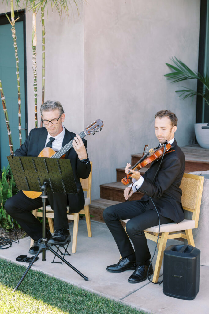 Malibu-Estate-Wedding_Malibu-Wedding_Southern-California-Wedding-Photographer_Lisa-Leanne-Photography_30-683x1024 Two men in suits sit on chairs outdoors, playing classical music instruments—a guitar and a violin—beside a modern building. Sheet music is on a stand in front of them, and a small speaker sits on the ground nearby. Copyright - Lisa Leanne Photography, California