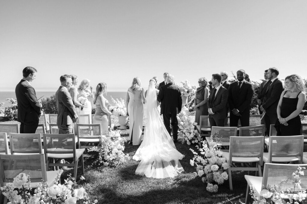 Malibu-Estate-Wedding_Malibu-Wedding_Southern-California-Wedding-Photographer_Lisa-Leanne-Photography_32-1024x683 A bride in a long veil walks down an outdoor aisle toward the groom, surrounded by guests seated and standing among floral arrangements, with an officiant waiting at the front; the scene is captured in black and white. Copyright - Lisa Leanne Photography, California