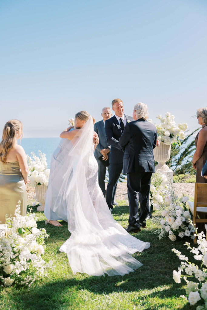 Malibu-Estate-Wedding_Malibu-Wedding_Southern-California-Wedding-Photographer_Lisa-Leanne-Photography_33-683x1024 A bride in a long white gown and veil hugs an older man outdoors at a seaside wedding ceremony, surrounded by white flowers, with guests and officiant watching on a sunny day. Copyright - Lisa Leanne Photography, California