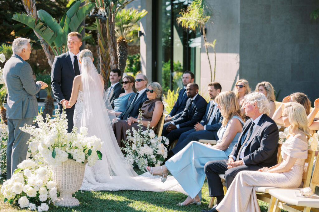 Malibu-Estate-Wedding_Malibu-Wedding_Southern-California-Wedding-Photographer_Lisa-Leanne-Photography_34-1024x683 An outdoor wedding ceremony with a bride in a veil and white gown standing beside the groom. Guests dressed formally are seated, surrounded by lush greenery and white floral arrangements under bright sunlight. Copyright - Lisa Leanne Photography, California