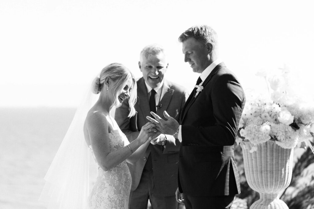 Malibu-Estate-Wedding_Malibu-Wedding_Southern-California-Wedding-Photographer_Lisa-Leanne-Photography_35-1024x683 A bride and groom exchange rings during their outdoor wedding ceremony, smiling as an officiant stands behind them. The scene is bright and joyful, with a floral arrangement visible on the right. Copyright - Lisa Leanne Photography, California