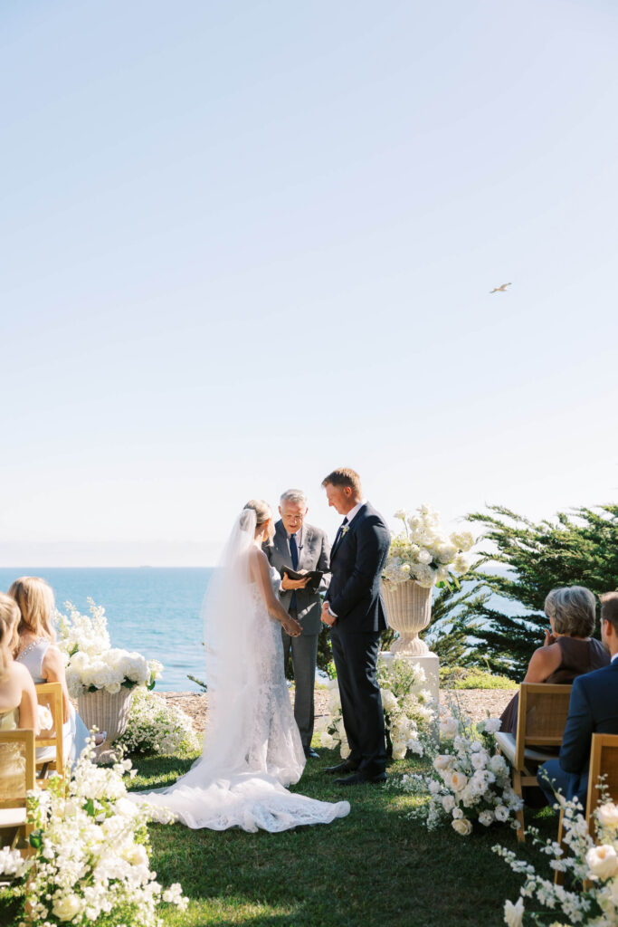 Malibu-Estate-Wedding_Malibu-Wedding_Southern-California-Wedding-Photographer_Lisa-Leanne-Photography_36-683x1024 A bride and groom stand facing each other during an outdoor wedding ceremony by the ocean, with a person officiating. Guests are seated on wooden chairs, and white flowers decorate the aisle and altar. Copyright - Lisa Leanne Photography, California