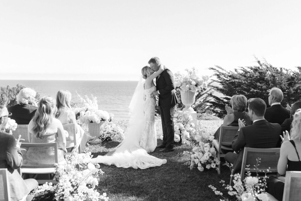Malibu-Estate-Wedding_Malibu-Wedding_Southern-California-Wedding-Photographer_Lisa-Leanne-Photography_37-1024x683 A bride and groom share a kiss at an outdoor wedding ceremony by the ocean, surrounded by guests seated on either side and floral arrangements decorating the aisle. Copyright - Lisa Leanne Photography, California