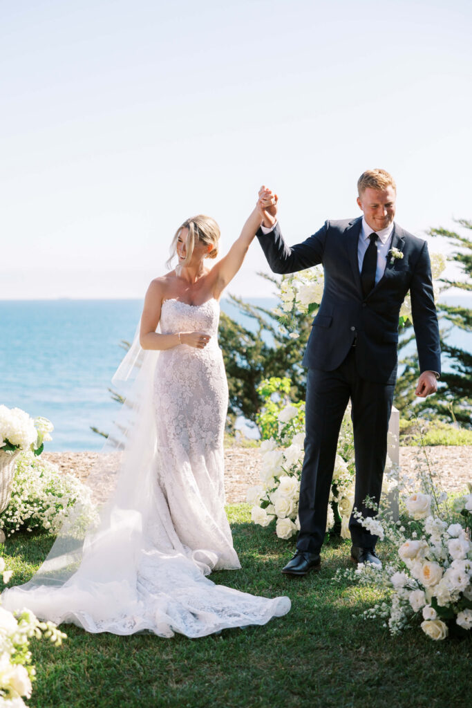 Malibu-Estate-Wedding_Malibu-Wedding_Southern-California-Wedding-Photographer_Lisa-Leanne-Photography_38-683x1024 A bride in a strapless white gown and a groom in a dark suit hold hands and celebrate outdoors, surrounded by white flowers, with the ocean and blue sky in the background. Copyright - Lisa Leanne Photography, California