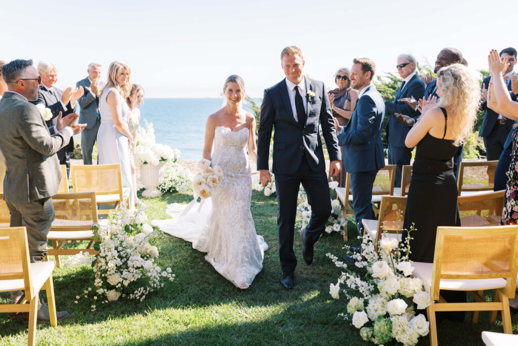 Malibu-Estate-Wedding_Malibu-Wedding_Southern-California-Wedding-Photographer_Lisa-Leanne-Photography_39-1024x683 A bride and groom walk down an outdoor aisle by the ocean, smiling and holding hands, as guests on either side stand, smile, and clap. The aisle is decorated with white flowers and wooden chairs. Copyright - Lisa Leanne Photography, California