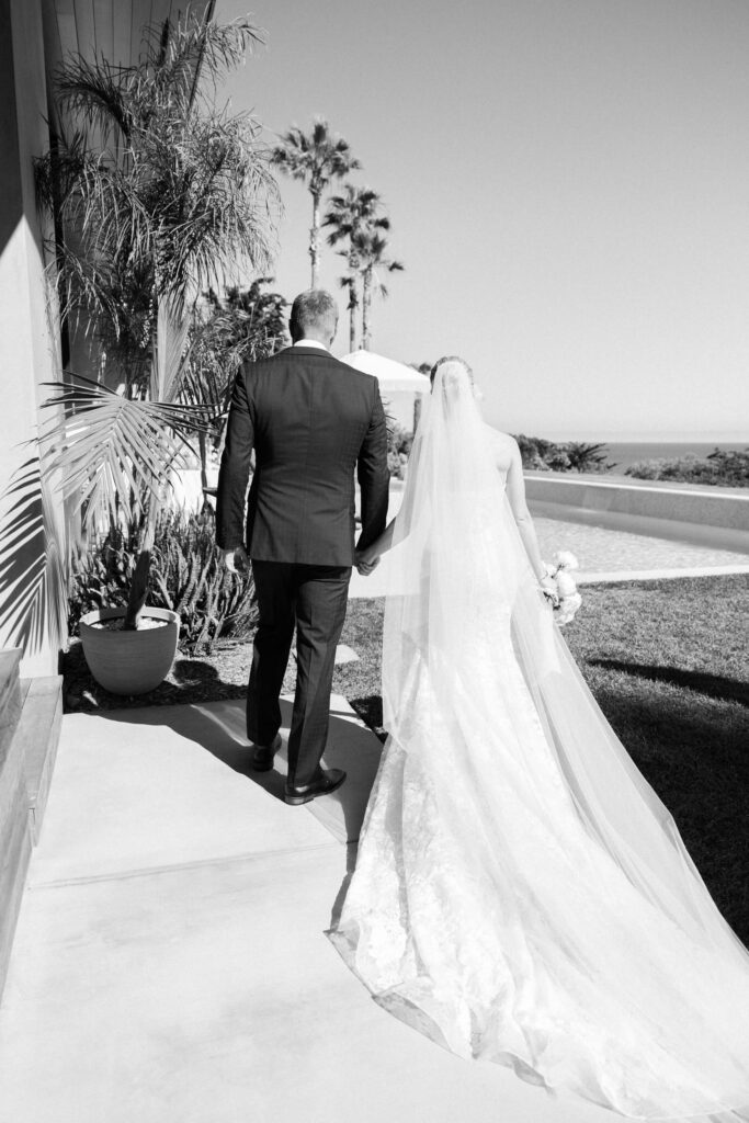Malibu-Estate-Wedding_Malibu-Wedding_Southern-California-Wedding-Photographer_Lisa-Leanne-Photography_40-683x1024 A bride and groom walk hand in hand outdoors, dressed in wedding attire. The bride wears a long gown with a veil, and the groom wears a suit. Palm trees and a sunny landscape are visible in the background. Copyright - Lisa Leanne Photography, California