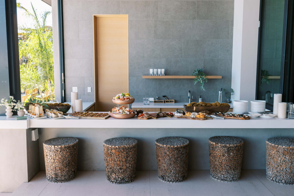 A modern buffet setup with assorted pastries and snacks on a white counter, five patterned stools in front, plates and glasses stacked to the right, and minimalist decor in a bright room with large windows. Copyright – Lisa Leanne Photography, California A modern buffet setup with assorted pastries and snacks on a white counter, five patterned stools in front, plates and glasses stacked to the right, and minimalist decor in a bright room with large windows. Copyright - Lisa Leanne Photography, California