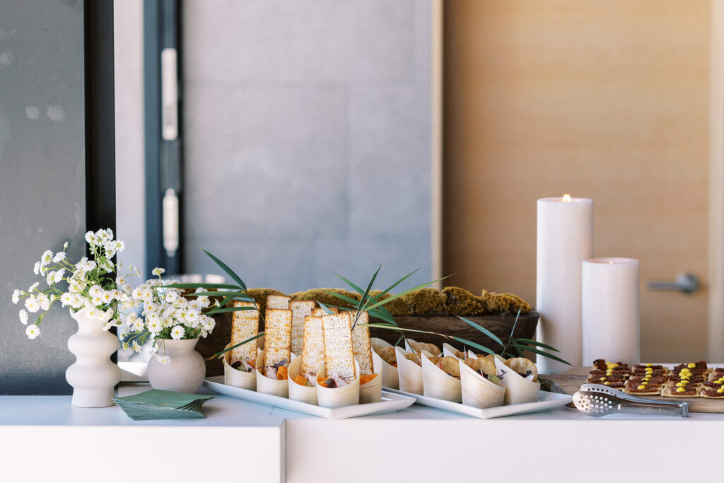 A modern white table set with matzo, small bowls of food, a plate of mini desserts, two white pillar candles, green napkins, a vase of white daisies, and greenery as decoration. Copyright – Lisa Leanne Photography, California A modern white table set with matzo, small bowls of food, a plate of mini desserts, two white pillar candles, green napkins, a vase of white daisies, and greenery as decoration. Copyright - Lisa Leanne Photography, California