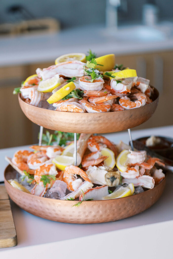 A two-tiered copper serving tray filled with cooked shrimp, crab claws, lemon wedges, and fresh herbs, displayed on a table. Copyright – Lisa Leanne Photography, California A two-tiered copper serving tray filled with cooked shrimp, crab claws, lemon wedges, and fresh herbs, displayed on a table. Copyright - Lisa Leanne Photography, California