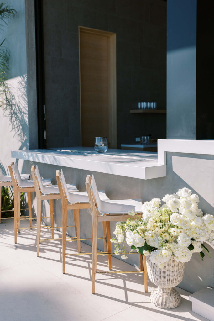 A modern outdoor bar area with wooden stools lined up at a white counter, a large vase filled with white flowers on the ground, and glassware atop the bar. Sunlight casts shadows on the light stone floor. Copyright – Lisa Leanne Photography, California A modern outdoor bar area with wooden stools lined up at a white counter, a large vase filled with white flowers on the ground, and glassware atop the bar. Sunlight casts shadows on the light stone floor. Copyright - Lisa Leanne Photography, California