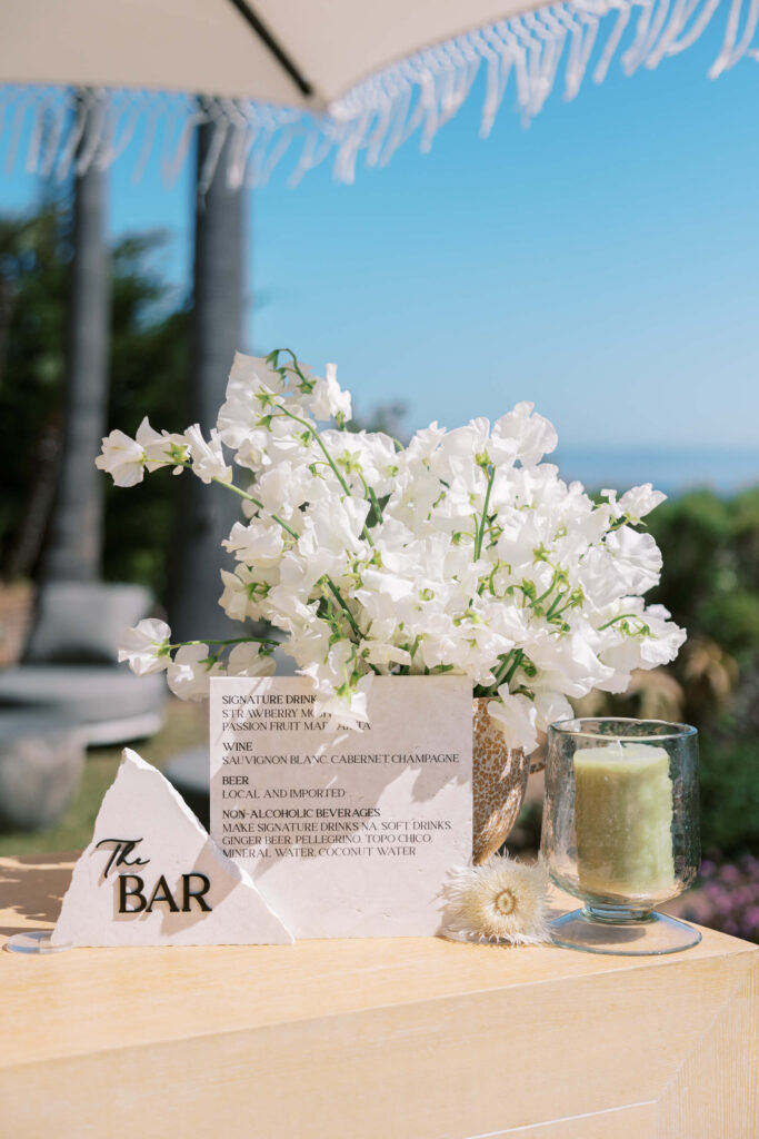 A bar menu on a table beside a green candle and a vase of white flowers, with palm trees and a blue sky in the background. The menu is printed on white paper and propped up with a white stone with “The Bar” written on it. Copyright – Lisa Leanne Photography, California A bar menu on a table beside a green candle and a vase of white flowers, with palm trees and a blue sky in the background. The menu is printed on white paper and propped up with a white stone with "The Bar" written on it. Copyright - Lisa Leanne Photography, California