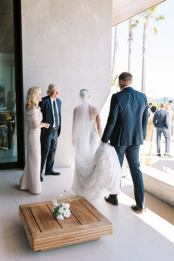 A bride in a white gown and veil walks with a man in a navy suit, while two elegantly dressed adults stand nearby. Sunlight streams in, and palm trees are visible outside through large windows. Copyright – Lisa Leanne Photography, California A bride in a white gown and veil walks with a man in a navy suit, while two elegantly dressed adults stand nearby. Sunlight streams in, and palm trees are visible outside through large windows. Copyright - Lisa Leanne Photography, California