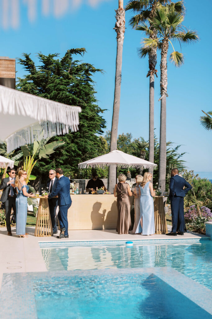People in formal attire gather and socialize around a poolside outdoor bar, surrounded by palm trees and greenery on a sunny day, with a blue sky in the background. Copyright – Lisa Leanne Photography, California People in formal attire gather and socialize around a poolside outdoor bar, surrounded by palm trees and greenery on a sunny day, with a blue sky in the background. Copyright - Lisa Leanne Photography, California