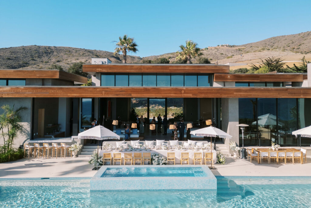 Malibu-Estate-Wedding_Malibu-Wedding_Southern-California-Wedding-Photographer_Lisa-Leanne-Photography_52-1024x683 A modern mansion with large glass windows and wooden accents features an outdoor pool. Elegant dining tables with white umbrellas and floral decorations are set up on the patio for an event, surrounded by hills and palm trees. Copyright - Lisa Leanne Photography, California