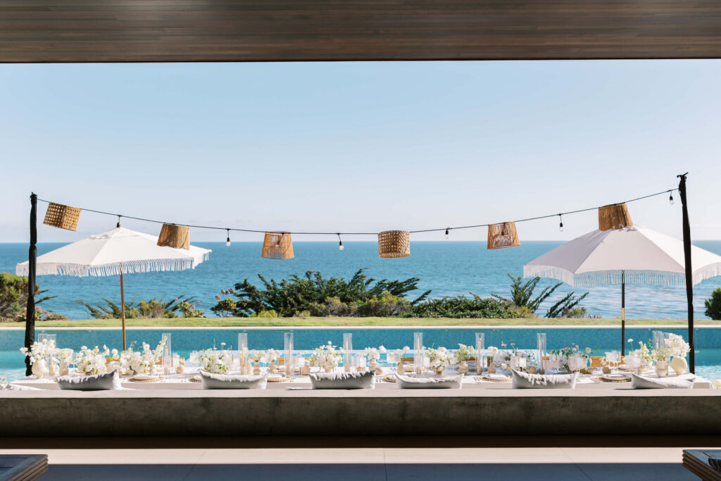 Malibu-Estate-Wedding_Malibu-Wedding_Southern-California-Wedding-Photographer_Lisa-Leanne-Photography_53-1024x683 A beautifully set outdoor dining table with white decor sits beside a pool, overlooking the ocean. Large umbrellas and string lights with woven lanterns provide shade and ambiance under a clear blue sky. Copyright - Lisa Leanne Photography, California
