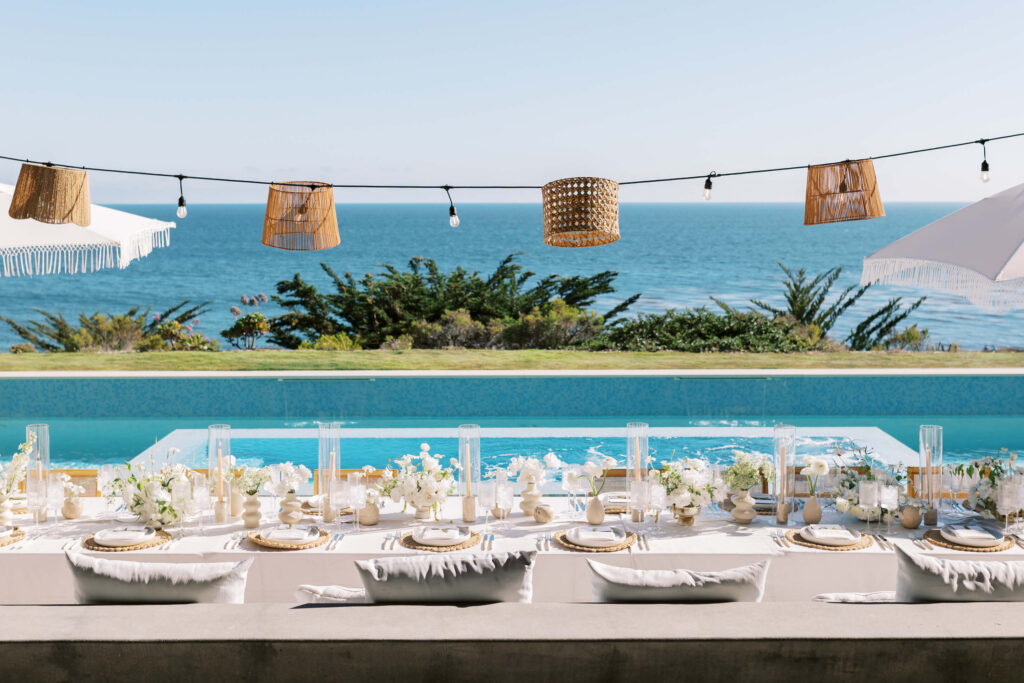 Malibu-Estate-Wedding_Malibu-Wedding_Southern-California-Wedding-Photographer_Lisa-Leanne-Photography_54-1024x683 A beautifully set outdoor dining table with white and gold decor overlooks a pool and the ocean. String lights with wicker lampshades hang above, and lush greenery and umbrellas frame the view. Copyright - Lisa Leanne Photography, California
