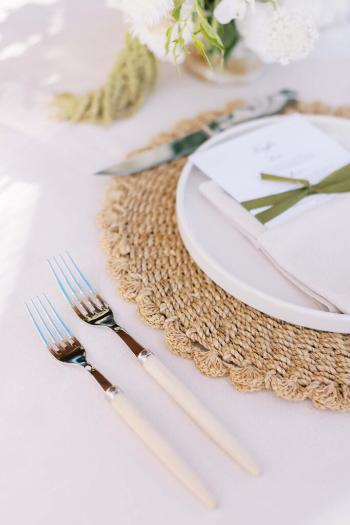 Malibu-Estate-Wedding_Malibu-Wedding_Southern-California-Wedding-Photographer_Lisa-Leanne-Photography_55-683x1024 A close-up of a table setting with two forks, a woven placemat, a white plate with a folded napkin and green ribbon, and a small bouquet of white flowers in the background. Copyright - Lisa Leanne Photography, California