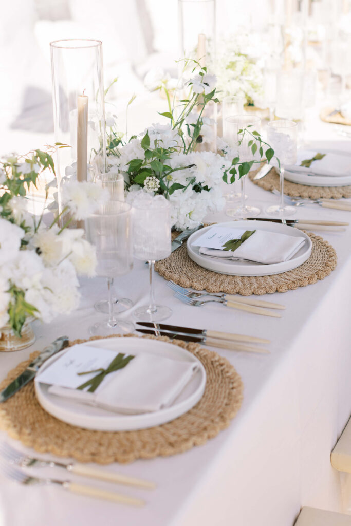Malibu-Estate-Wedding_Malibu-Wedding_Southern-California-Wedding-Photographer_Lisa-Leanne-Photography_56-683x1024 Elegant table setting with white plates, linen napkins tied with green leaves, woven placemats, glassware, candles, and white floral centerpieces on a white tablecloth. Bright, airy atmosphere. Copyright - Lisa Leanne Photography, California