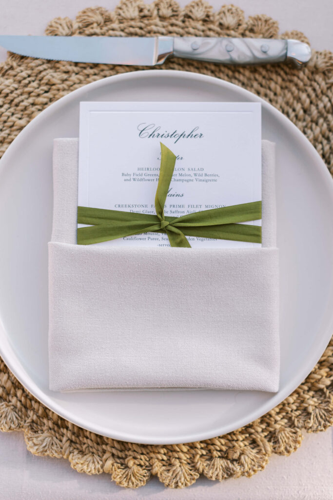 Malibu-Estate-Wedding_Malibu-Wedding_Southern-California-Wedding-Photographer_Lisa-Leanne-Photography_57-683x1024 A white plate on a woven placemat holds a neatly folded white napkin, with a printed menu card tied with a green ribbon tucked inside. A knife with a light handle rests above the plate. Copyright - Lisa Leanne Photography, California