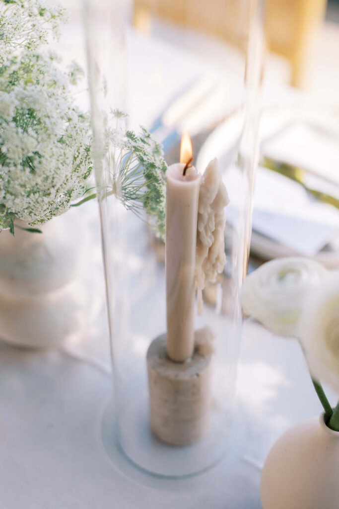 Malibu-Estate-Wedding_Malibu-Wedding_Southern-California-Wedding-Photographer_Lisa-Leanne-Photography_58-683x1024 A lit taper candle with melted wax drips stands in a stone holder inside a clear glass cylinder, surrounded by delicate white flowers on a softly lit table. Copyright - Lisa Leanne Photography, California